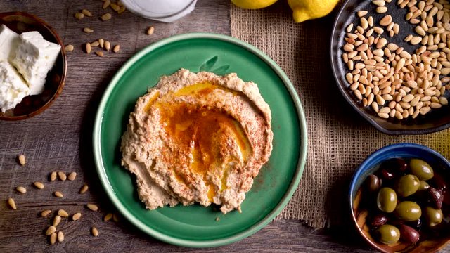 Wide sliding to the left showing a flat lay plate of hummus with feta, olives, glass of ouzo, lemons, pine nuts on a rustic wood table.