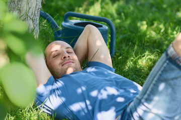 Man in blue t-shirt lying on grass with hands behind head in the garden with watering can nearby on blurred greenery background on a sunny summer day. Free time and shying away from work concept