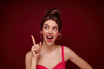 Attractive excited young brown haired female with evening makeup wearing her brown hair in bun and showing upwards positively with raised forefinger, posing over burgundy background