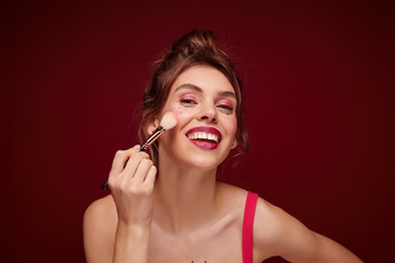 Indoor shot of beautiful young brown haired lady with casual hairstyle looking positively in mirror and applying makeup with brush, preparing for going out, isolated over burgundy background