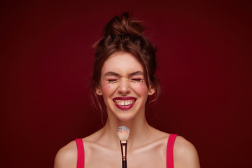 Pleasant looking cheerful young brunette lady with bun hairstyle and evening makeup rejoicing about upcoming party and smiling happily with closed eyes, isolated over burgundy background