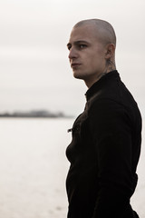 Bald attractive man in tattoos on the ocean. Tattooed brutal young man stands on the ocean in a black denim jacket. A stern guy posing while on the coastline by the sea. Stylish and fashionable guy.
