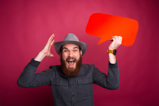 Shocked young bearded hipster man holding speech bubble and gesturing