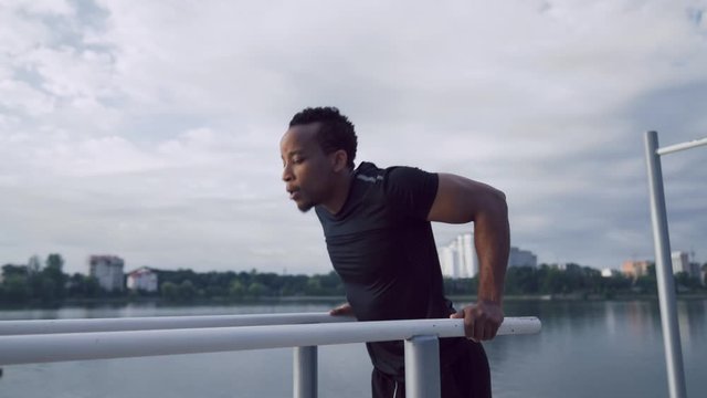 Afro american guy workout on dips bars near the city lake