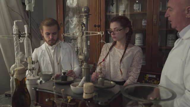 Close-up of young man and woman listening to mature pharmacist in white robe and holding test tubes. Two new workers in ancient drugstore learning medicine. Vintage pharmacy, retro, 19th century.