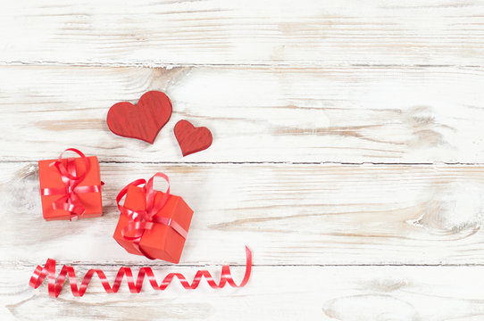 two red wooden hearts and two red gift boxes with bows underneath a red shiny twisted ribbon at the bottom left of the image on a white wooden background top view