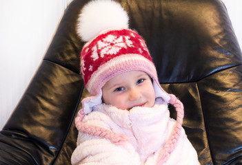 A girl in a knitted winter hat with a pumpkin sits on a black, leather armchair. Baby in a warm bathrobe