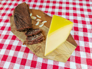 Smoked beef meat sliced with garlic cloves on the side with a block of hard yellow cheese on a wooden cutting board with red colorful tablecloth