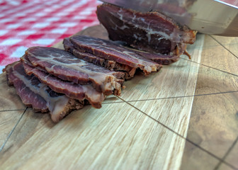 A man cutting smoked beef meat with a knife on a wooden cutting board with slices stacking and red and white tablecloth in the background
