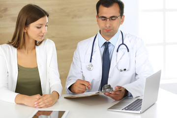 Doctor and patient discussing the results of a physical examination while sitting at a desk in a clinic. A male doctor using a clipboard to fill out a medical history of a young woman's medication