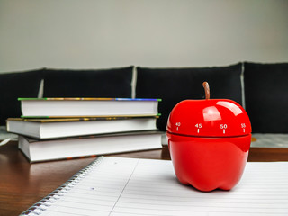 Pomodoro study technique with apple shaped kitchen timer standing on a white empty notebook and books stacked in the background on a wooden desk