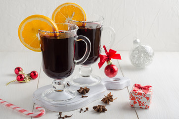 Two glass glasses of mulled red wine with orange slices stand on a white wooden table. On the table are Christmas decorations(red balls, sugar cane and a small box with a gift).