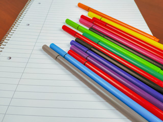 Fototapeta premium Closeup of many pens with different colors laying on the top of the white empty notebook with lines and spiral for note taking in school and better organization