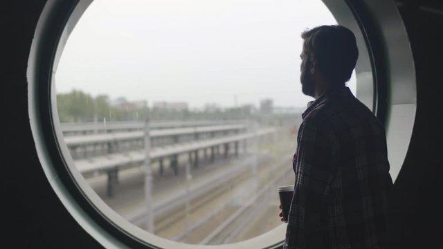 Male young traveler at train station looking out round window and drinking coffee.
