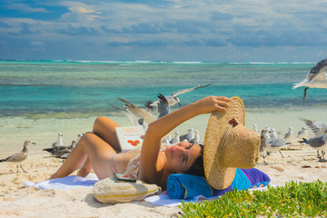 caucasian woman lying on sand beach, reading a book with screened Laughing gull flying around her. Caribbean destination