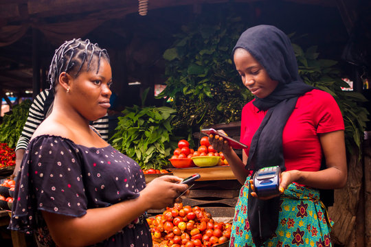 African Woman Shopping Food Stuff In A Local Market Paying By Doing Mobile Transfer Via Phone For A Trader