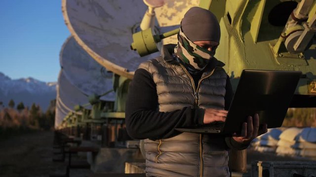 Hacker hacks the radio telescope using notebook. Man to program hacking on laptop in nature. Terrorist is hiding in camouflage balaclava. Hacker has criminal cases on the Internet. Programmer uses a