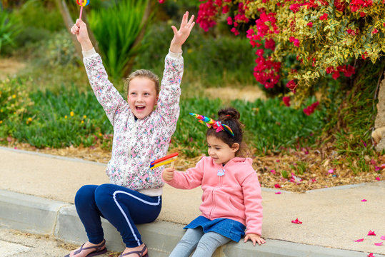 Funny teenager girl holding wooden grogger and happy toddler girl dressed as unicorns with noisemaker have fun outdoor. Purim, carnival, birthday party concept.