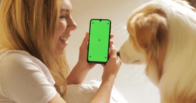 GREEN SCREEN tracking dot. Portrait of Pretty girl student is using smartphone shows screen and laughing lying on bed at home with cute pet dog border collie , animal is enjoying care and love.