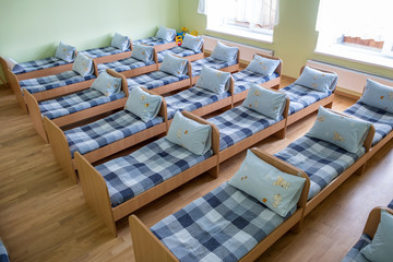 Many small beds with fresh linen in daycare preschool empty bedroom interior for comfortable afternoon nap of the kids. - obrazy, fototapety, plakaty