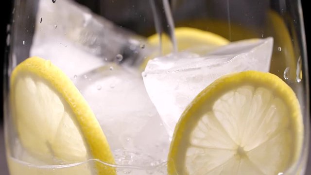 Pouring of soda water into glass with ice and lemon, closeup. Slow motion effect