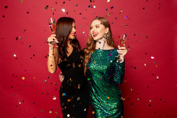 two beautiful models, a redhead and a brunette in New Year's dresses, having fun and smiling with glasses of champagne, confetti flying around on a red background. New Year or Christmas photo