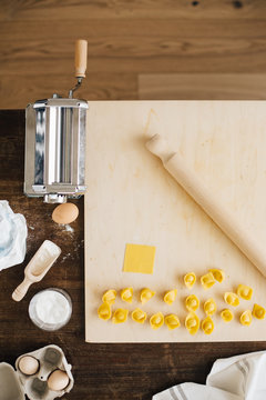 Freshly Made Ravioli Cappelletti on wooden Cutting Board with rolling pin, pasta maker and ingredients seen from above