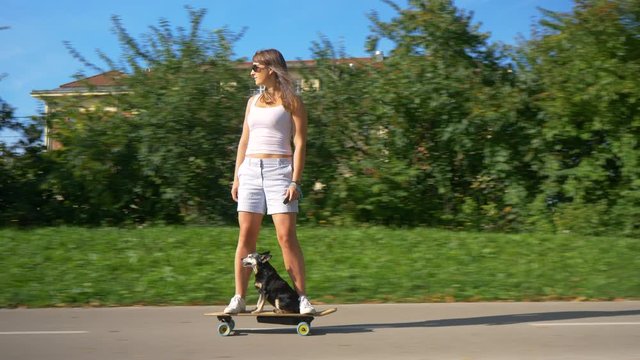 Happy millennial girl and her cute little miniature pinscher cruise through the scenic park on a cool e-longboard. Young Caucasian woman skateboards with her adorable senior dog on a sunny summer day.