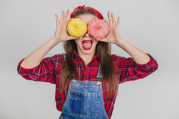 Lovely young female hiding her eyes behind two big yellow and pink doughnuts, making faces, showing her tongue.