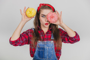 Childish young girl is hiding her eye behind big doughnut with pink icing, holding yellow doughnut near head, making duck face.
