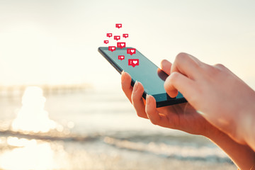 Concept of social networks. Female hands hold a smartphone and press something in it. Icons of likes fly over the screen. At the rear of the palne sea and sunset. Close up