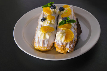 Two cakes with cream and fresh fruits on a white plate. The cakes are decorated with blueberry, mandarin slices and mint leaves.