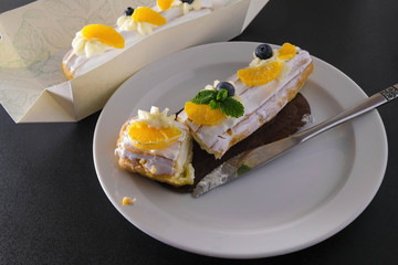 Two cakes with cream and fresh fruits. Blueberry, tangerine slices and mint leaves. Cut pastry with a knife on a white plate. Another cake in a box.