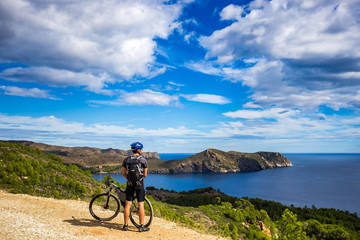 a young guy on a mountain bike trails in Spain and takes a photo on a white phone in the background of the Mediterranean sea of the rocky coast of the Costa Brava - obrazy, fototapety, plakaty