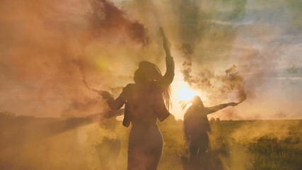 Naklejka na ściany i meble Happy friends girls waving colorful smoke at sunset.
