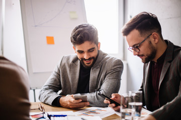 Two young good-looking programmers using their smart-phones while they are waiting for team meeting in the conference room to start.