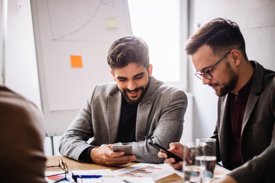 Two young good-looking programmers using their smart-phones while they are waiting for team meeting in the conference room to start.