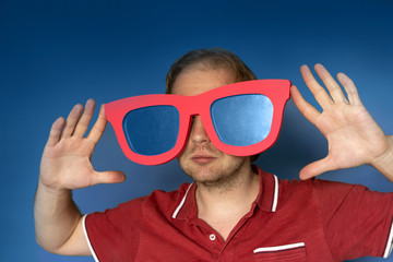 A fair-haired man in a classic red polo shirt holding big toy glasses with blue glasses in his hands in front of his eyes. The concept of exaggeration, another reality, vision, blind