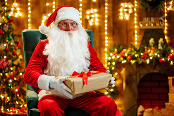 Photo of happy Santa Claus in eyeglasses sitting on chair, hold gift box and looking at camera
