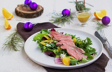 Warm salad with a mixture of salad leaves, baked duck and orange on a clay plate on a light concrete background in the New Year or Christmas style. Salad recipes with duck. Holiday recipes.