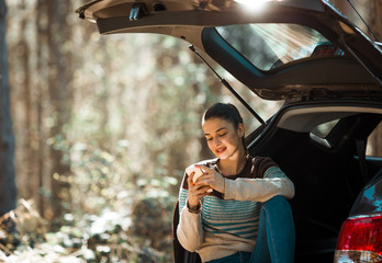 A woman sitting in a car truck, using her phone, with her elbow resting on her knee. The picture is taken in nature with blurred row of trees in the background.