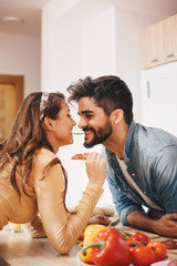 A man and a woman moments before kissing over the kitchen countertop. The woman is holding a carrot. Vegetables are layed on the counter as well.