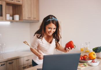 A woman watching her favorite TV show while preparing a tasty meal in the kitchen. She is holding a spatula and a red bell pepper.