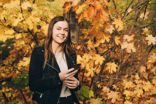 Portrait of smiling teenage girl with mobile phone standing against trees during autumn