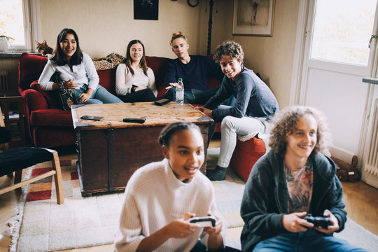Smiling male and female playing video games while friends sitting in background at home