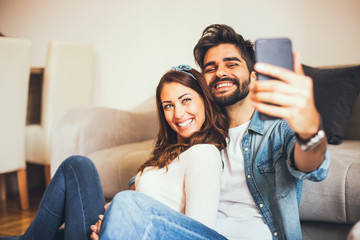 Young Caucasian couple smiling and taking a selfie while they are embracing and sitting by the sofa in their living room.