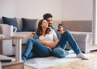 Fototapeta premium Young attractive Caucasian couple embracing and sitting on the floor by the sofa in the living room and watching photographs on smart-phone.