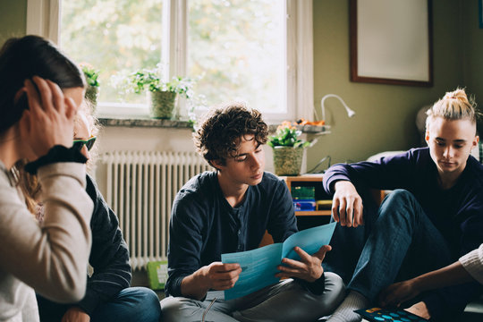Teenage boy reading brochure while sitting with friends in bedroom