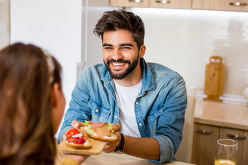 Fototapeta na wymiar Handsome bearded Caucasian man giving his girlfriend a healthy sandwich while they are sitting in the kitchen.