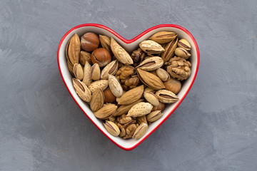 Variety of nuts in red bowl heart shaped on gray background. almonds, walnuts, hazelnuts, pistachios. healthy food for the heart. top view. horizontal